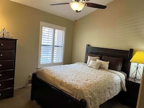 A bedroom with a bed, a dresser, a nightstand with a lamp, a ceiling fan, and a window with blinds.