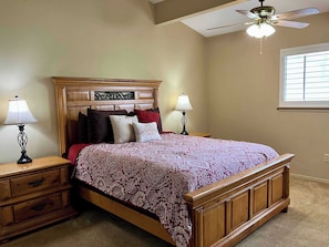 A bedroom with a wooden bed, two bedside tables with lamps, a ceiling fan, and a window with blinds. - Golf Course Front Condo | Ruidoso Getaway w/ (Ruidoso)