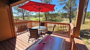 A wooden deck with a table and chairs, a red umbrella, and a view of trees and hills. - Grisham’s View Condo| Cozy w/Golf Course Views! (Ruidoso)