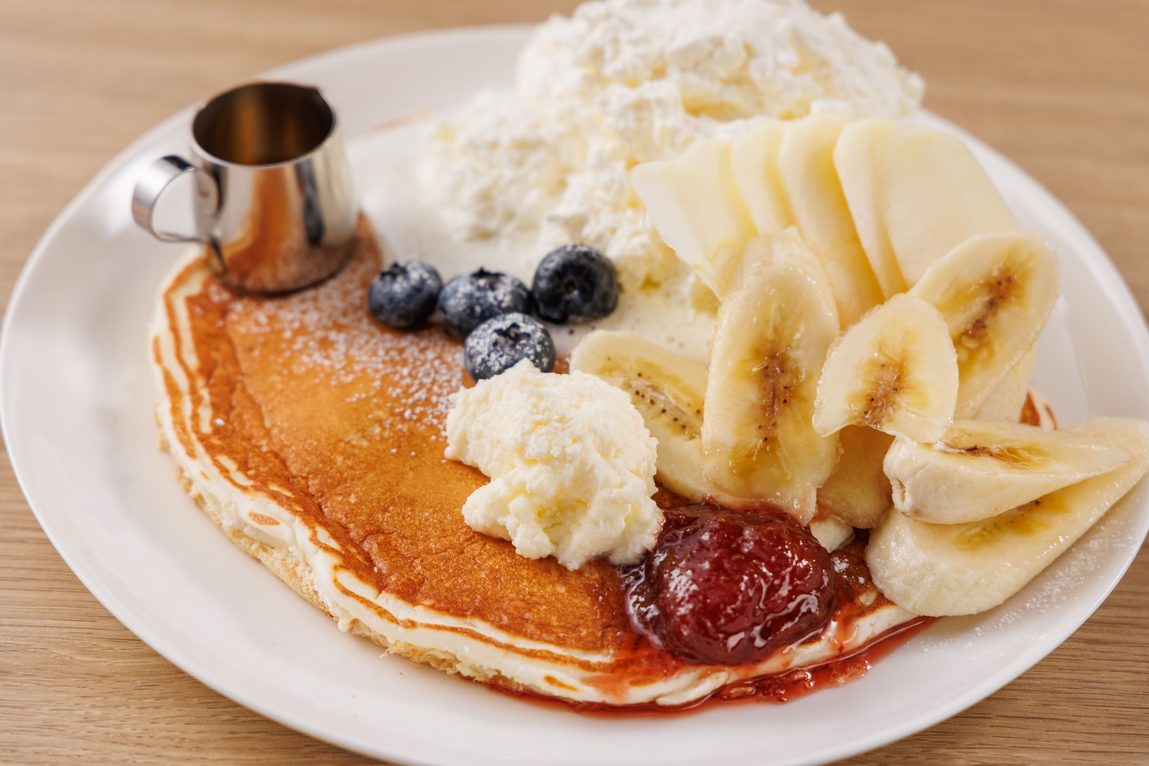 A plate of pancakes with bananas, blueberries, and a small metal cup.