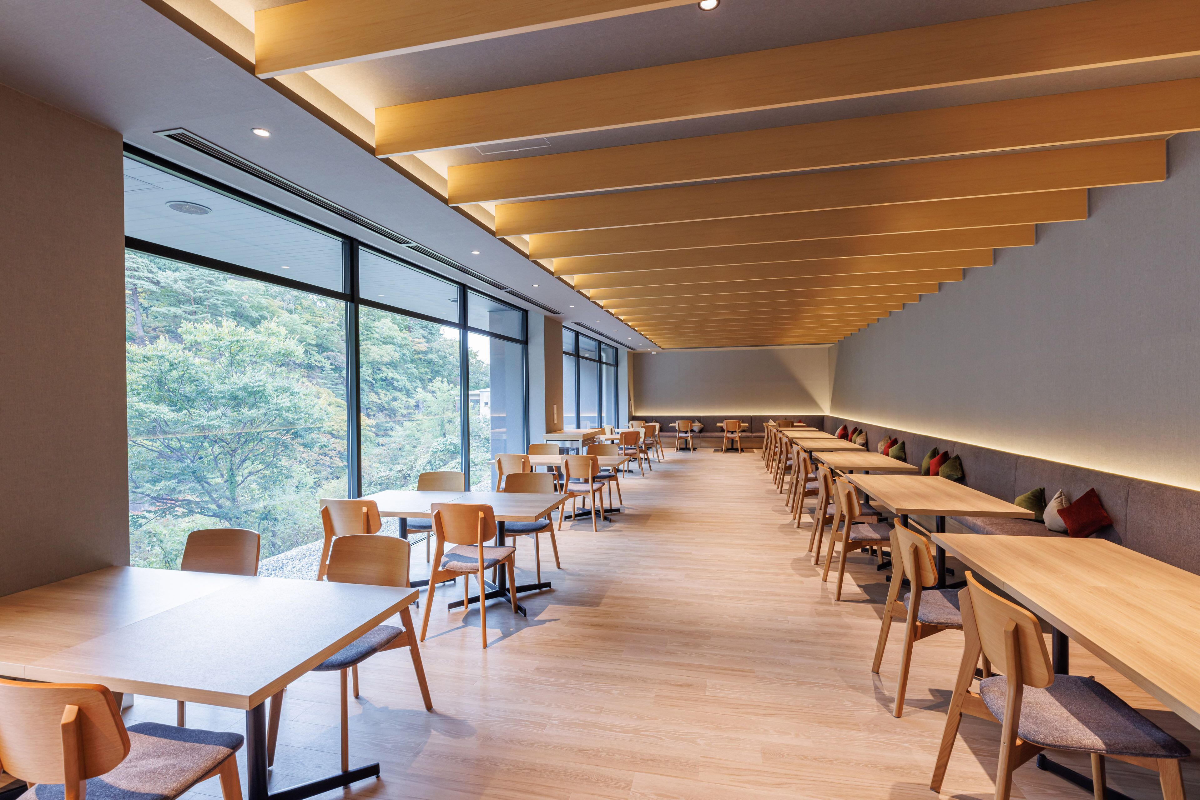 A modern dining area with long wooden tables and chairs, large windows offering a view of trees, and a sloped ceiling with recessed lighting.
