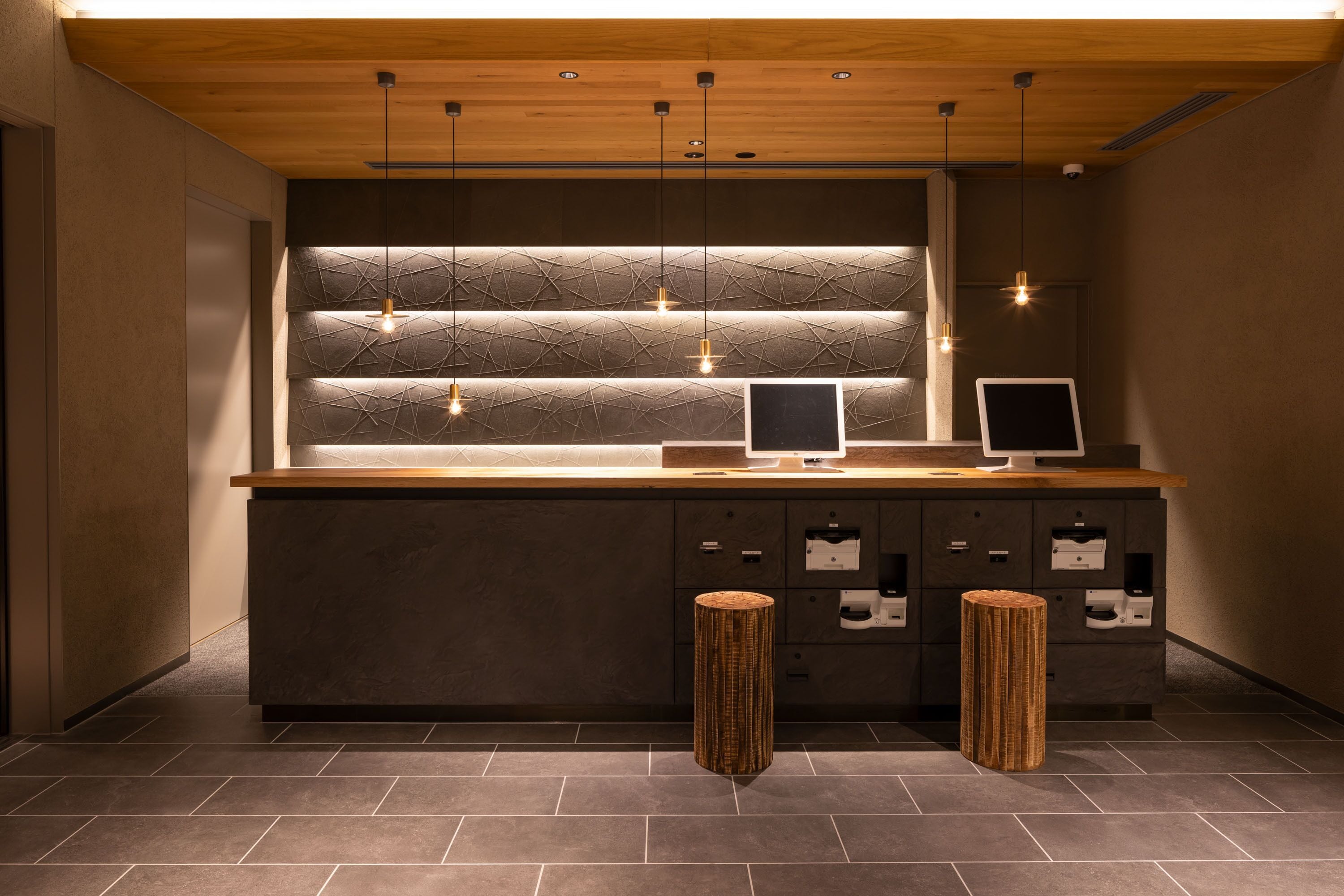 A modern reception desk with a wooden top, two monitors, and a textured wall behind. There are two woven stools in front.