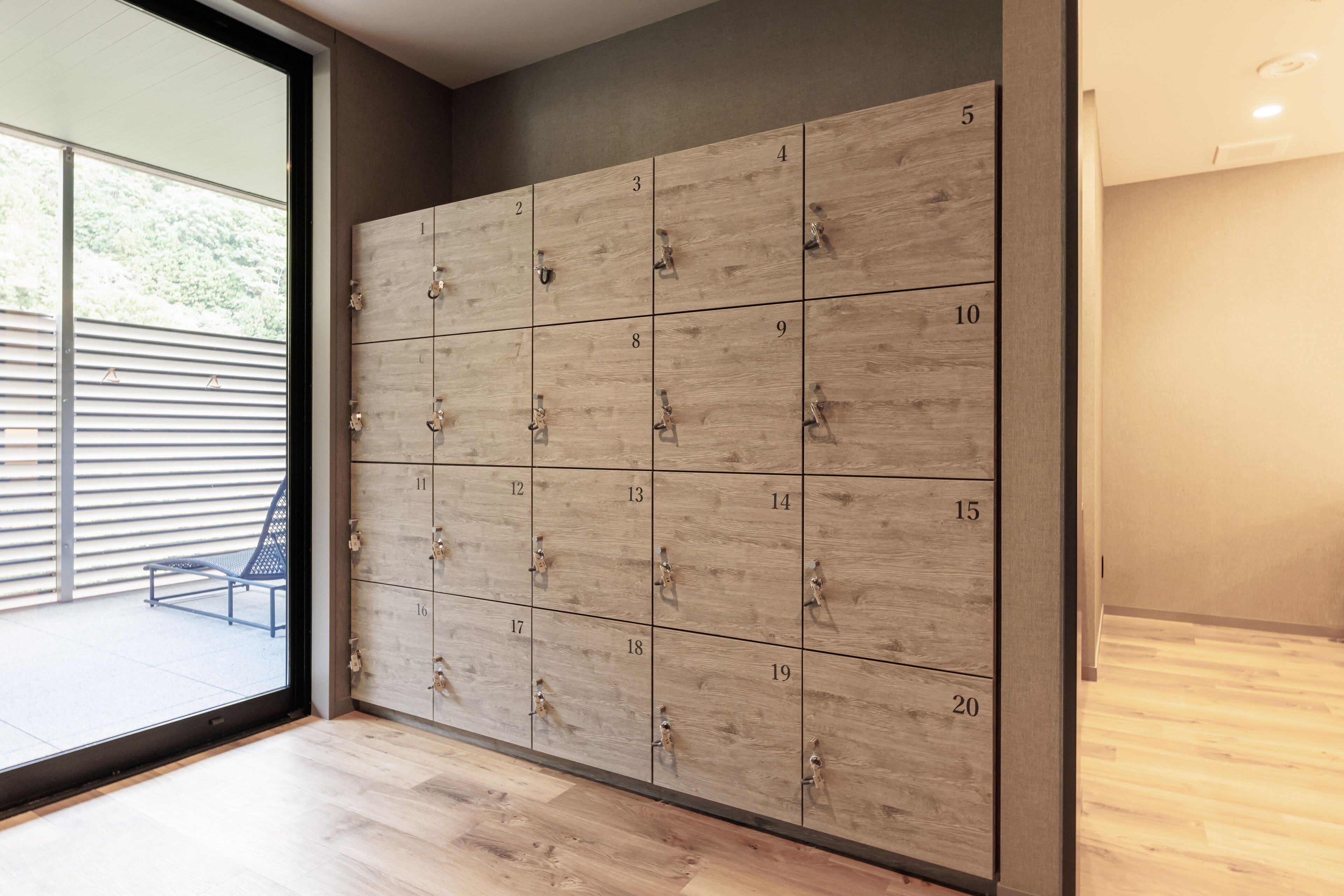 A row of wooden lockers with numbers from 1 to 20, a bench outside, and a view of a building through a glass door.