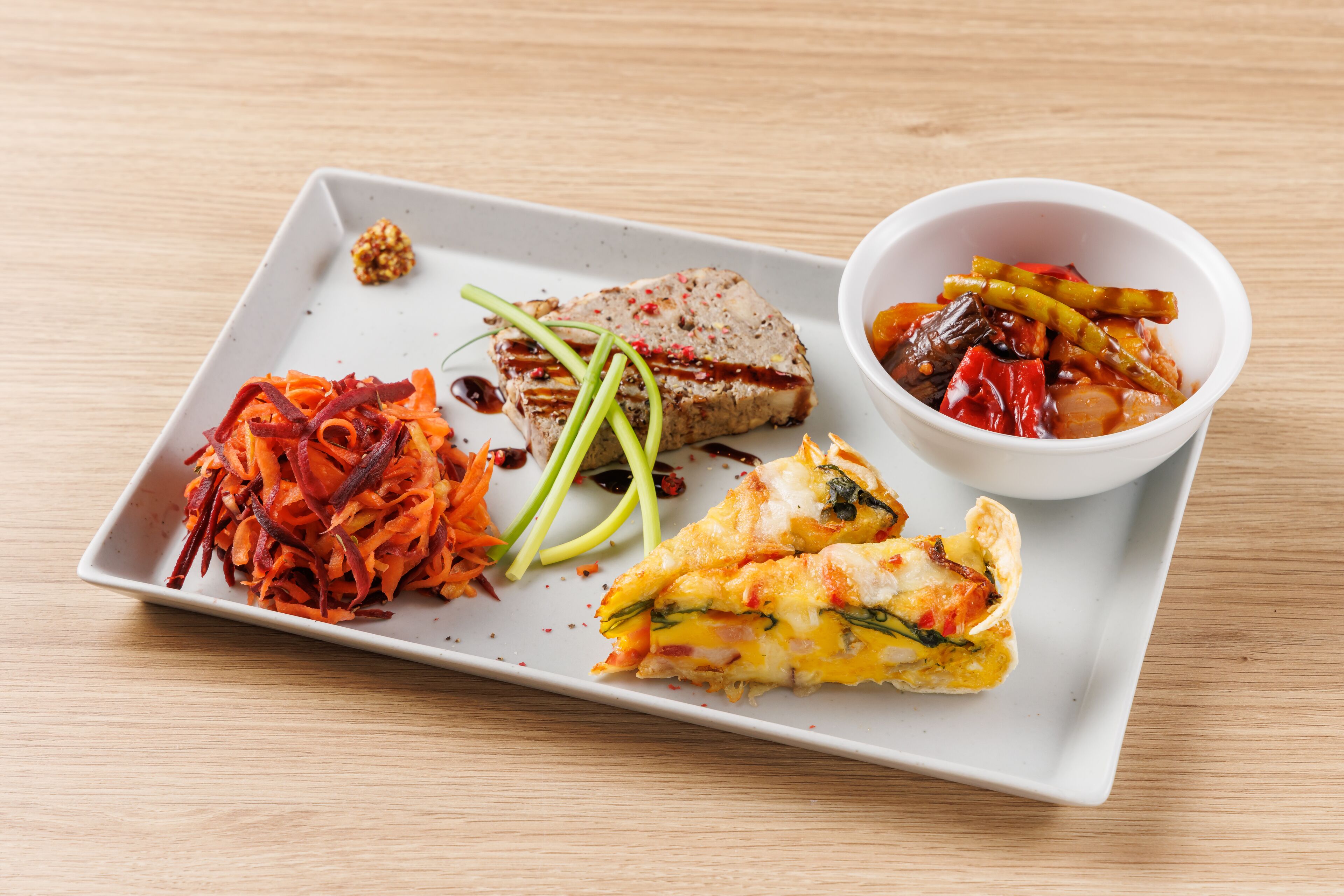Une assiette rectangulaire blanche contenant divers aliments, notamment un morceau de viande, un plat de légumes et une tranche d’omelette.