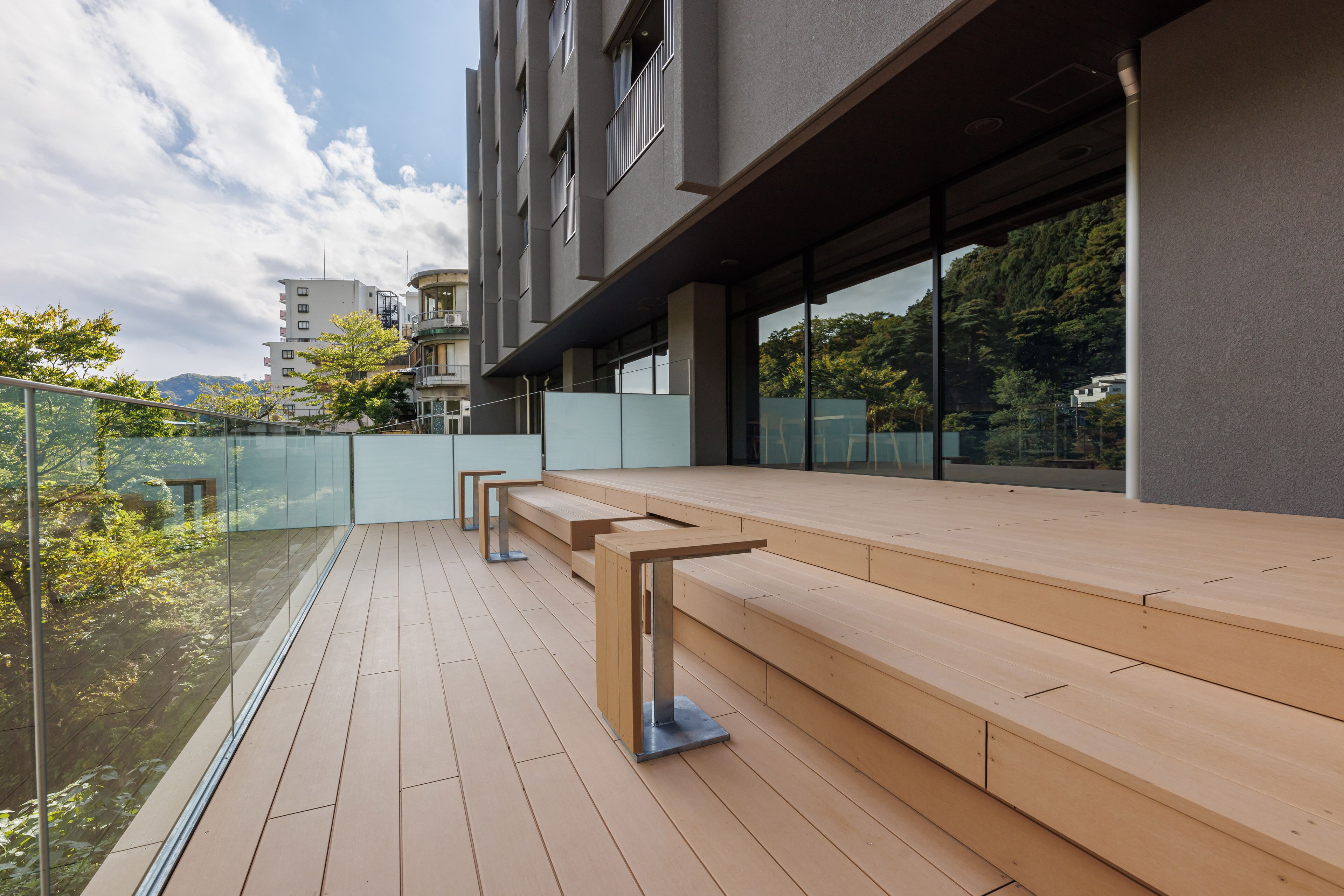 A modern outdoor seating area with wooden benches and glass railings, overlooking a cityscape and greenery.