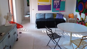 Una sala moderna con sofá, mesa de comedor y una silla. Hay un ventilador, un aparador y un florero con flores.