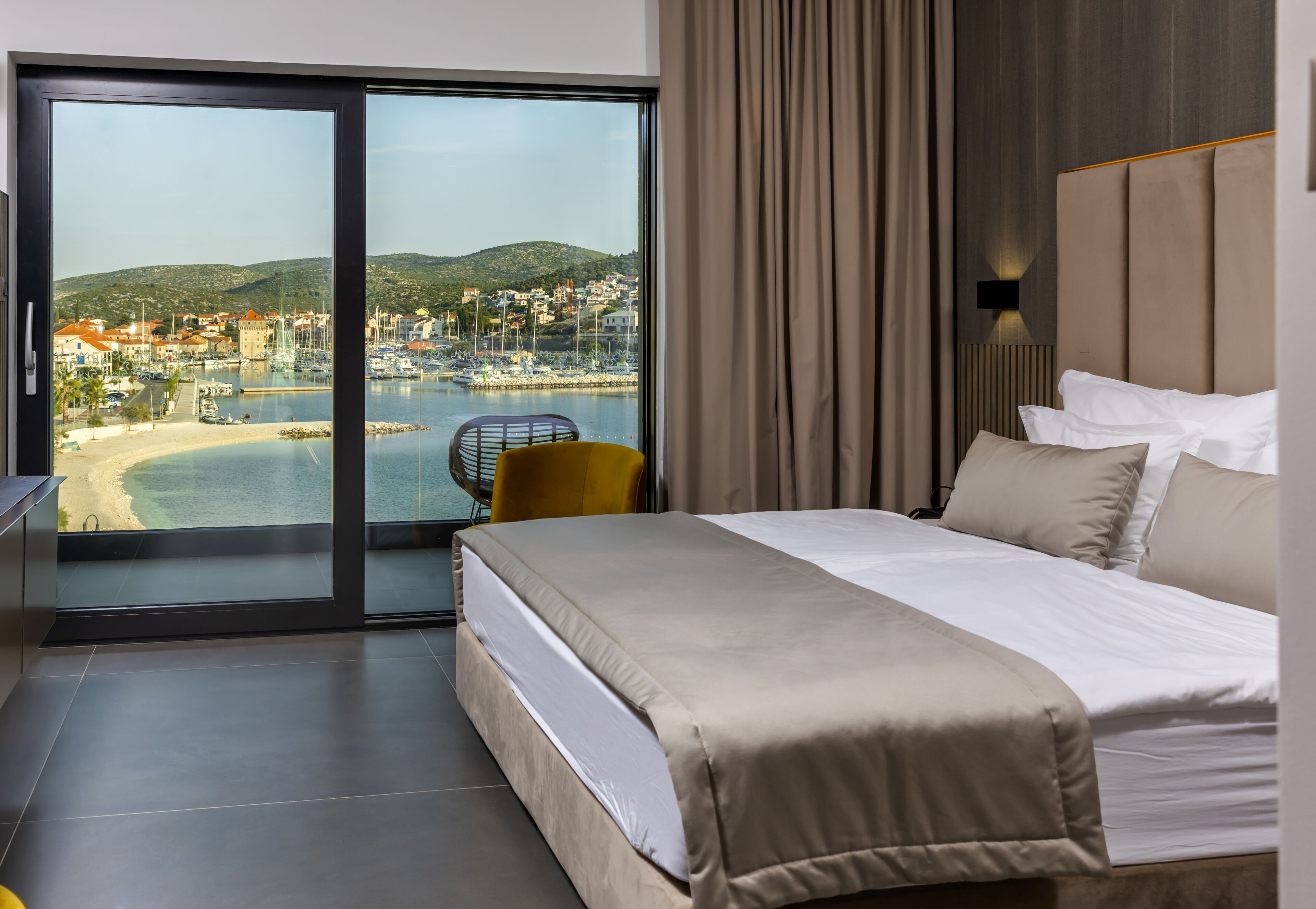 A hotel room with a bed, a chair, a view of a coastal town, and a large window.