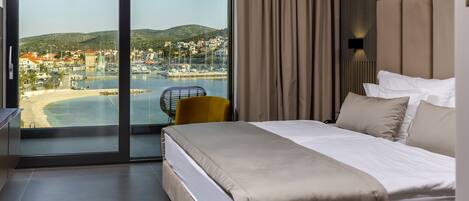 A hotel room with a bed, a chair, a view of a coastal town, and a large window.