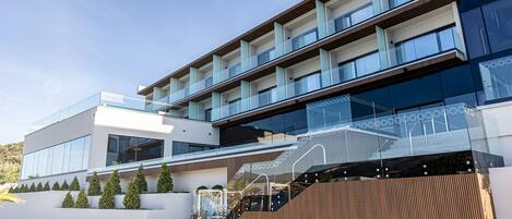 A modern multi-story hotel with a glass facade, multiple balconies, and a landscaped garden area.
