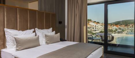A modern hotel room with a large bed, a view of a coastal town, and a balcony with outdoor seating.