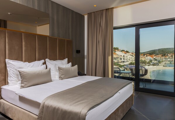 A modern hotel room with a large bed, a view of a coastal town, and a balcony with outdoor seating.
