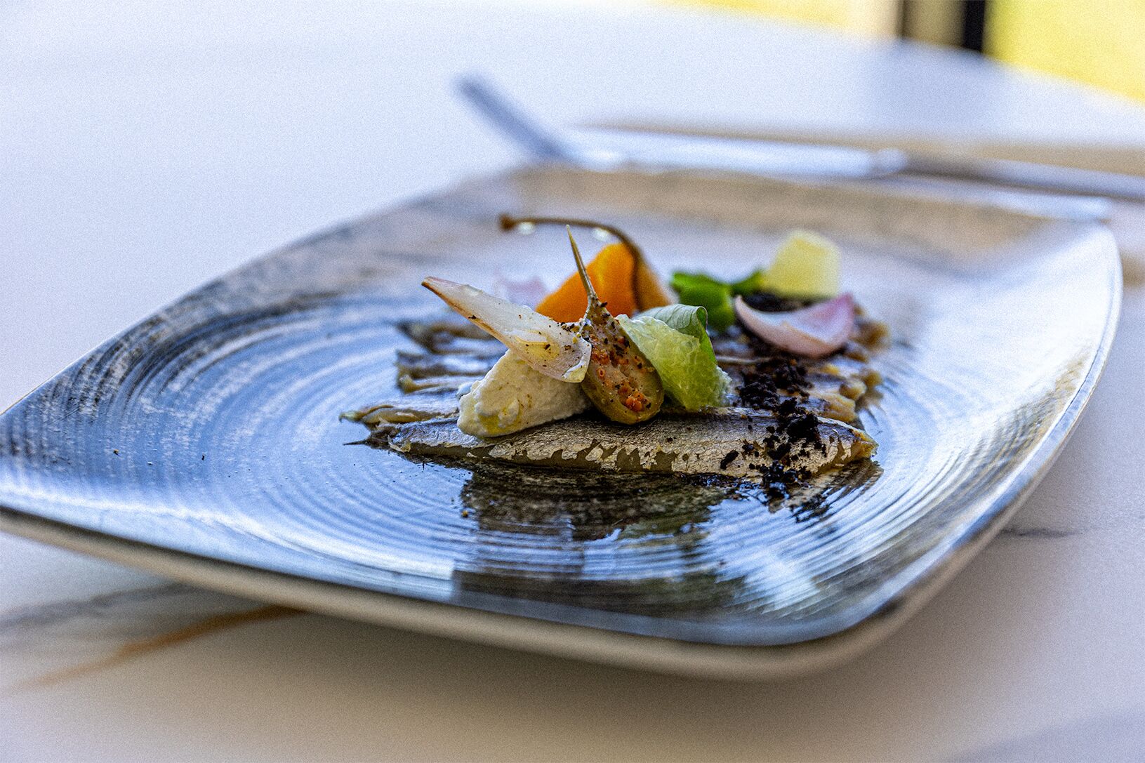 A square plate with a seafood dish, including fish and vegetables, on a table.