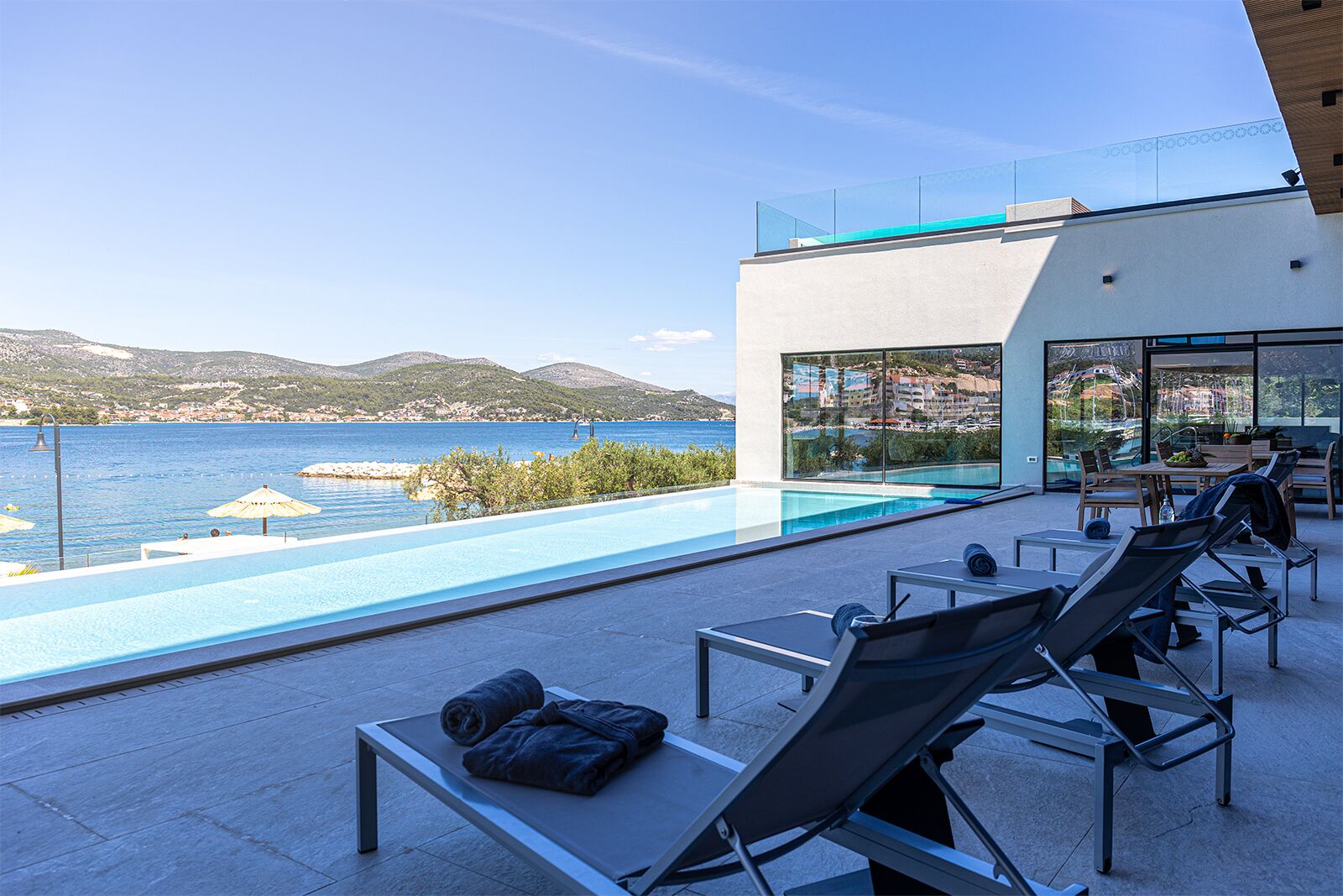 A rooftop pool with lounge chairs and a table, overlooking a coastal view with mountains in the distance.
