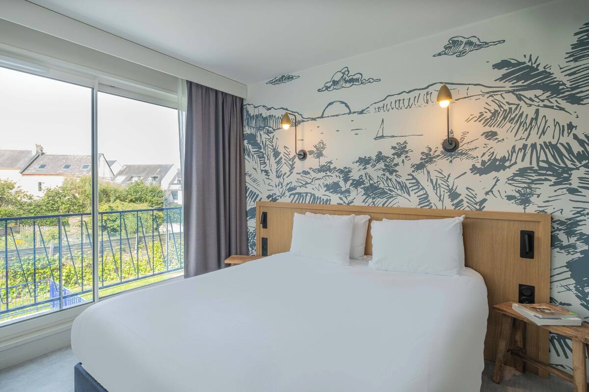 Ein Hotelzimmer mit einem großen Bett, einem Fenster mit Blick auf Gebäude und einem Wandbild.
