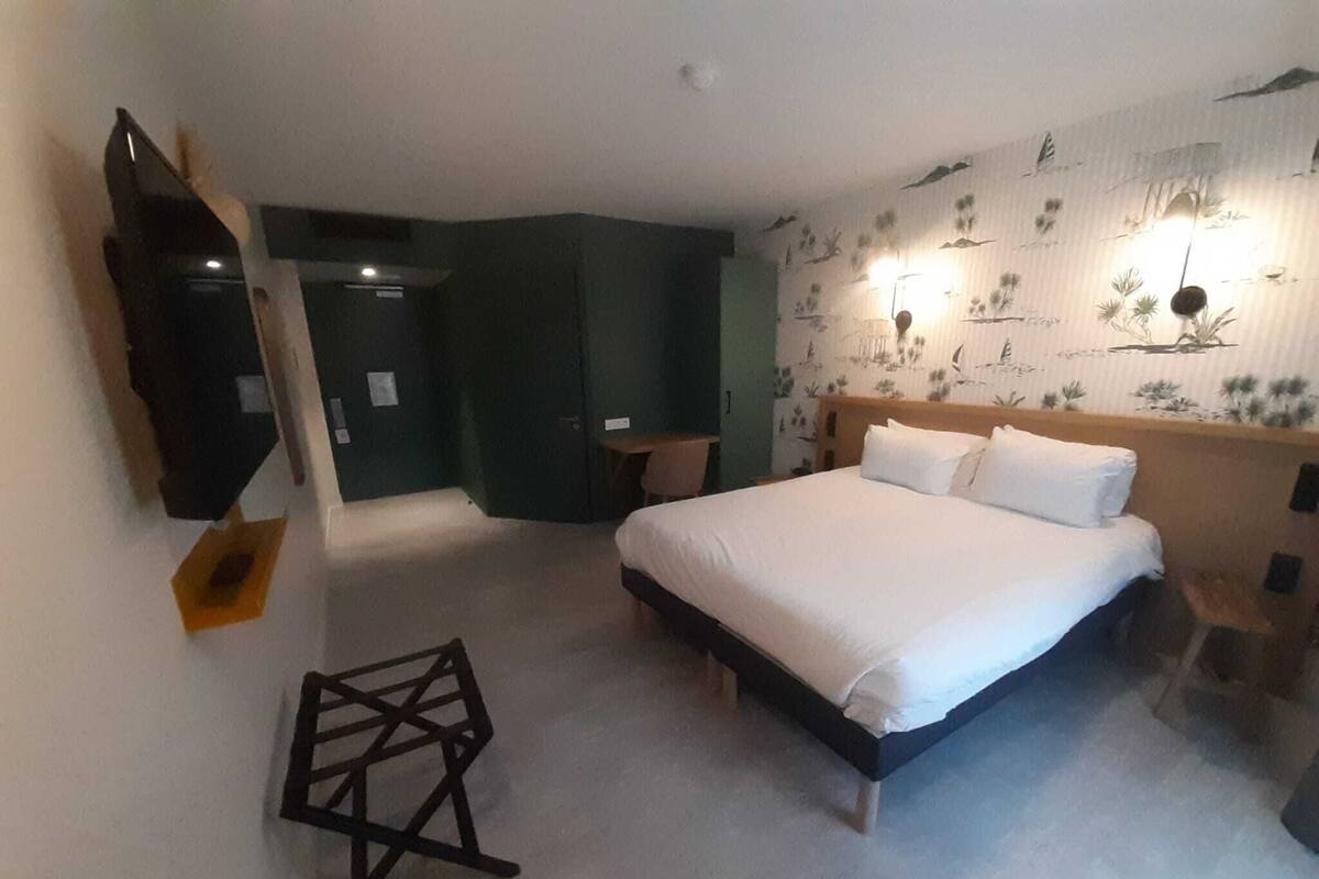 Ein Hotelzimmer mit Bett, Schreibtisch, Sessel und einem an der Wand befestigten Fernseher.