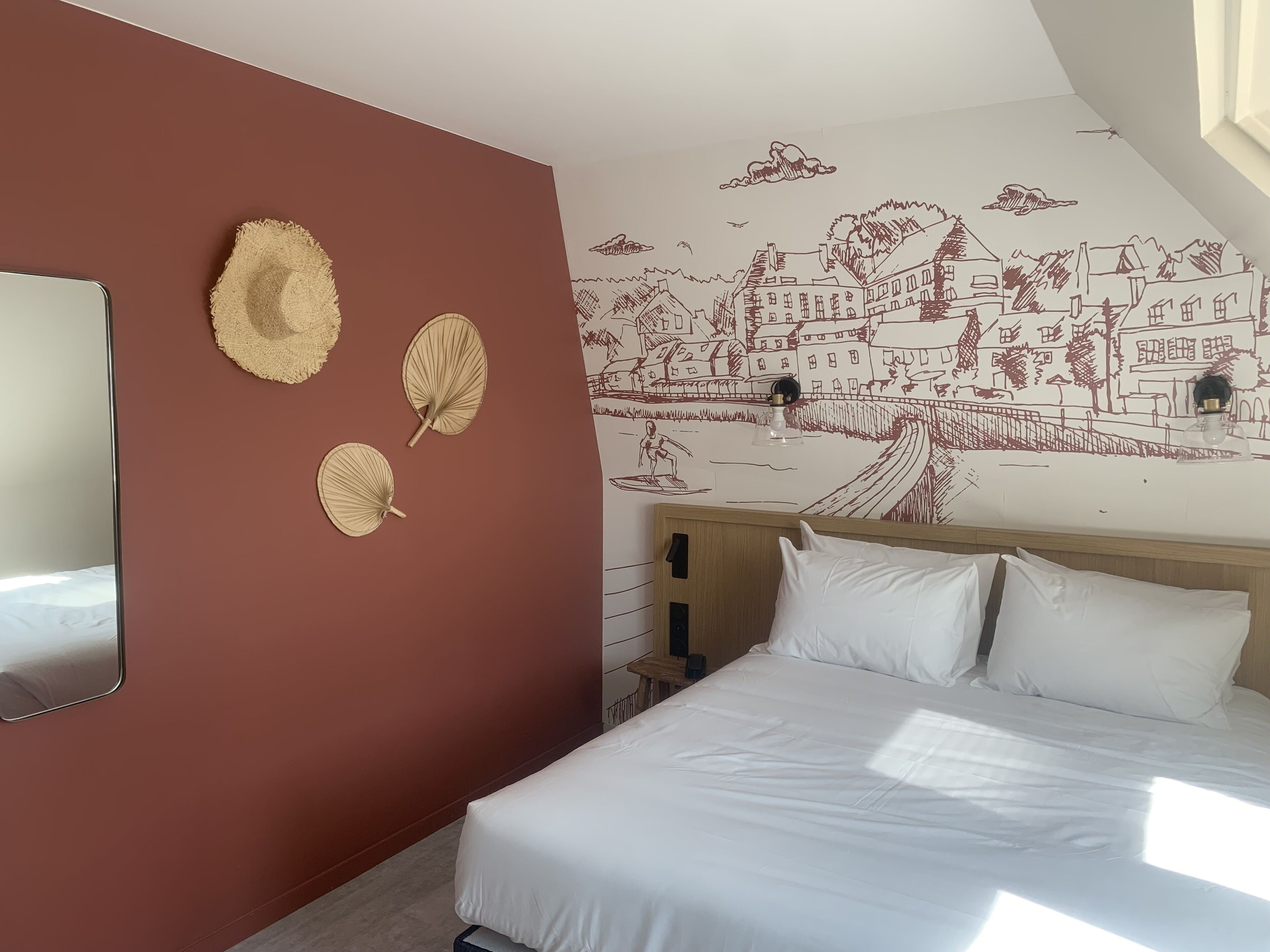 A bedroom with a bed, a wall-mounted mirror, and a mural of a town scene.