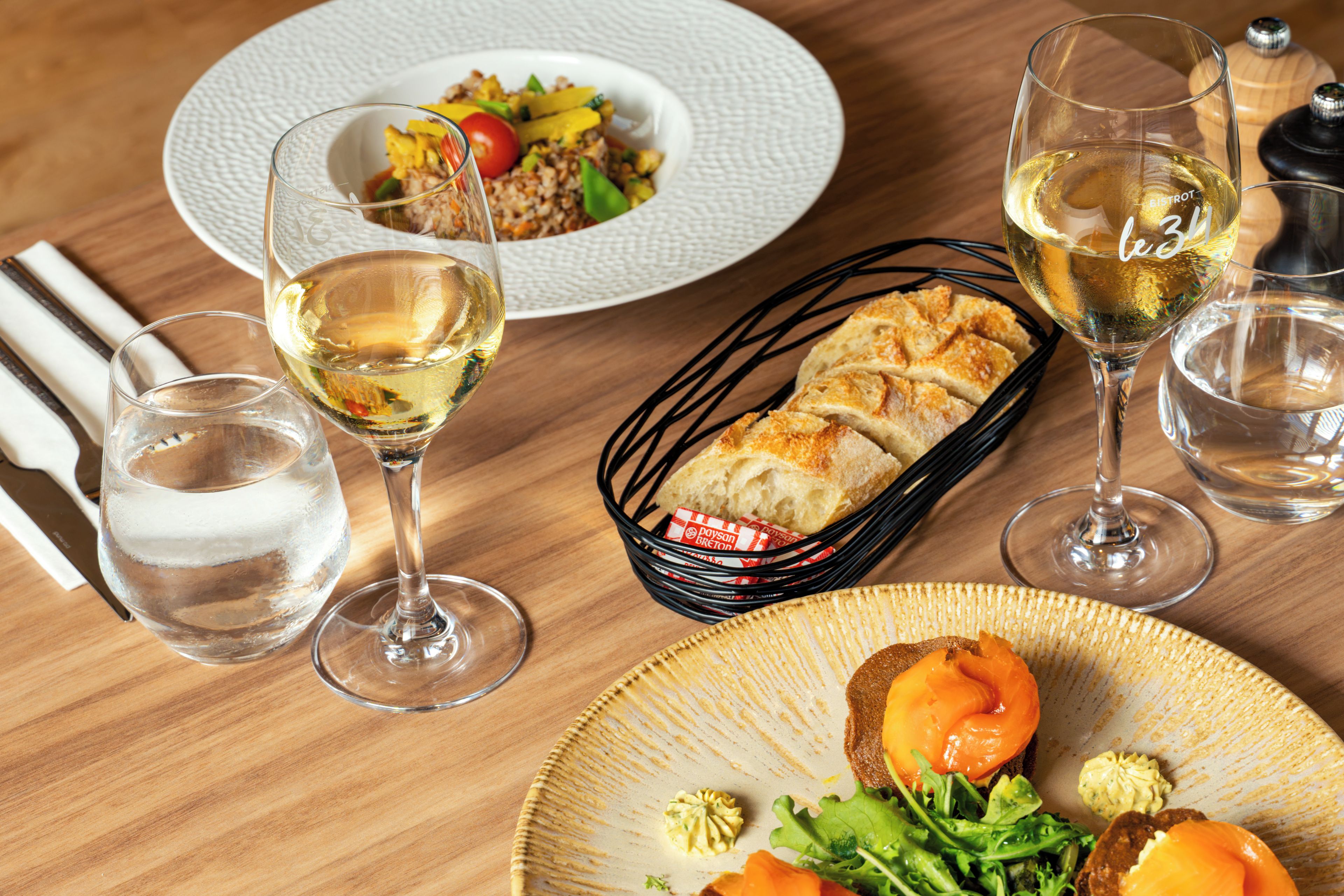 A dining table with a variety of dishes, including a salad, bread, and a plate with salmon and greens. There are also several glasses of white wine.
