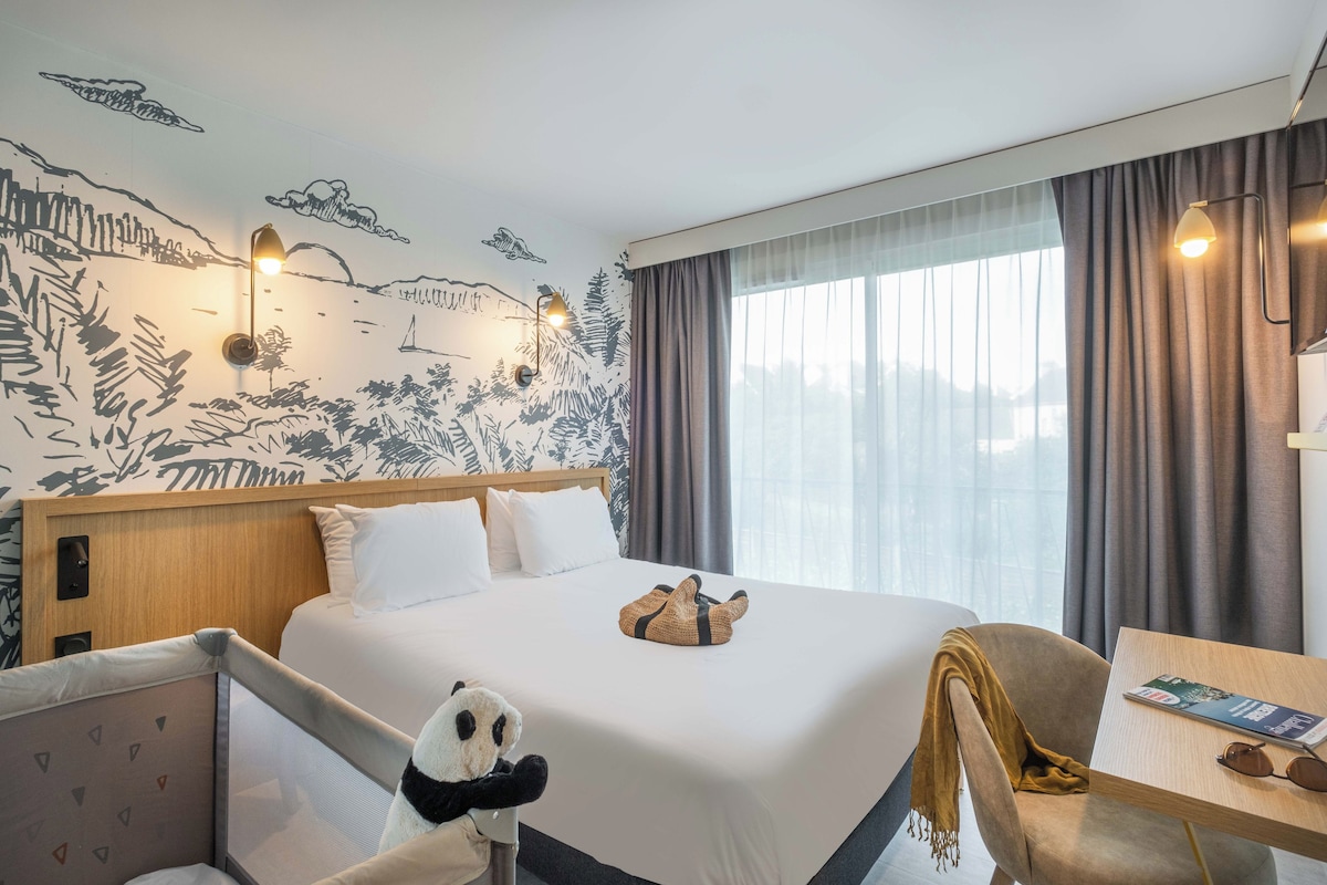 A hotel room with a bed, a desk, a panda plush toy, and a mural on the wall.