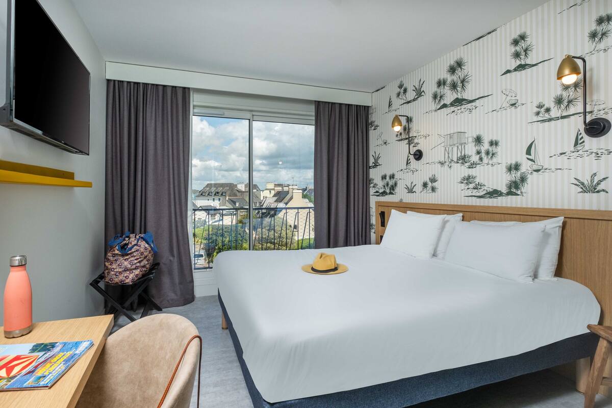 Ein Hotelzimmer mit einem großen Bett, einem Schreibtisch mit Stuhl, einem an der Wand montierten Fernseher und einem Fenster mit Vorhängen.