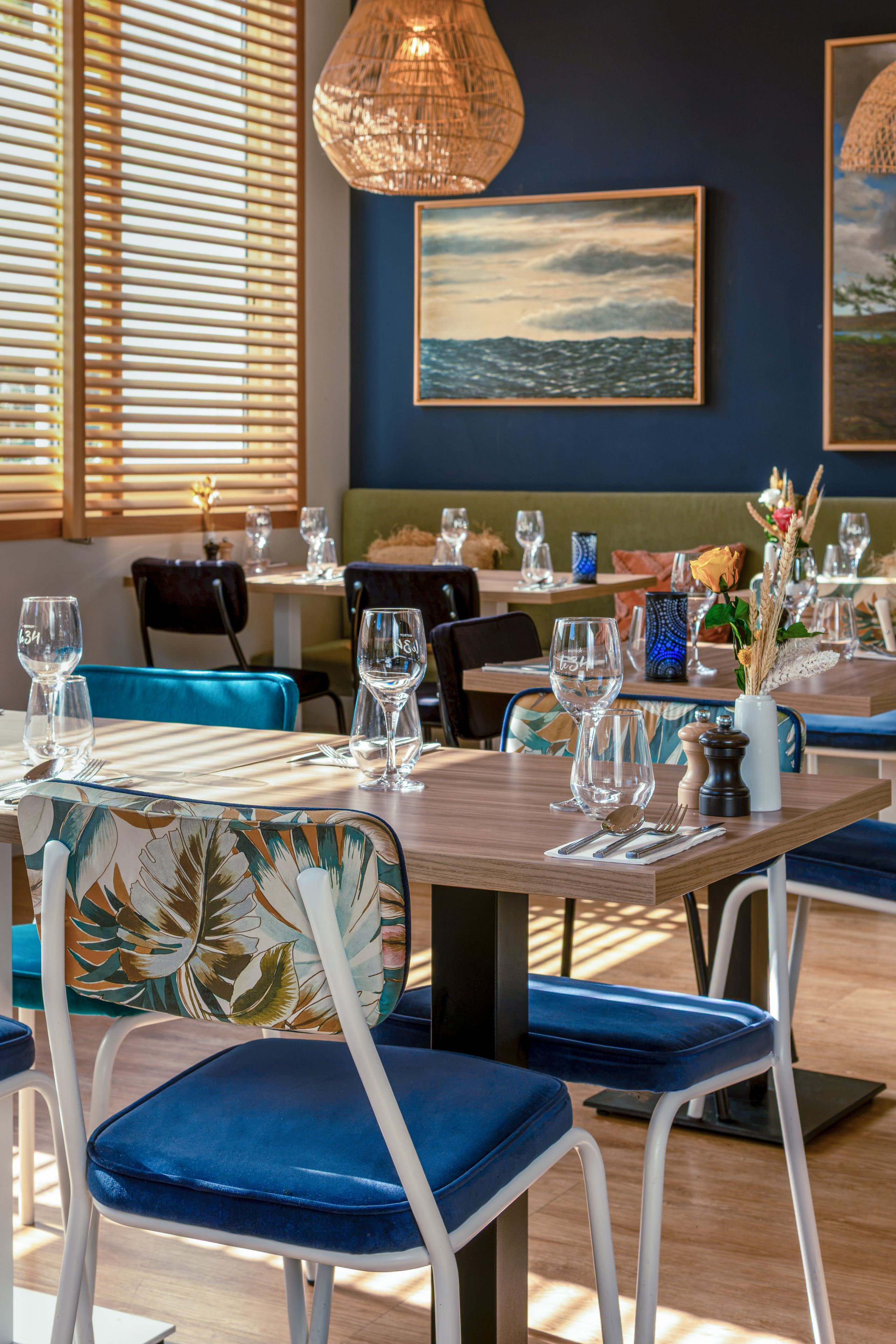 Un interior de restaurante con mesas dispuestas para comer, sillas con cojines azules y un cuadro de un paisaje marino en la pared.