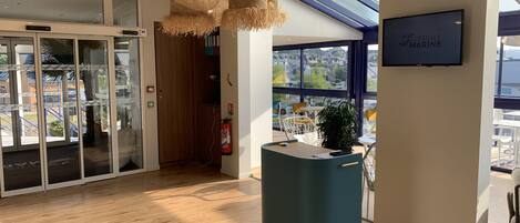 A modern interior with a reception desk, hanging wicker lamps, a fire extinguisher, and a large window offering a view of the outside.