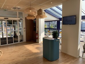 Un interno moderno con una reception, lampade di vimini a sospensione, un estintore e un'ampia finestra che offre vista all'esterno.