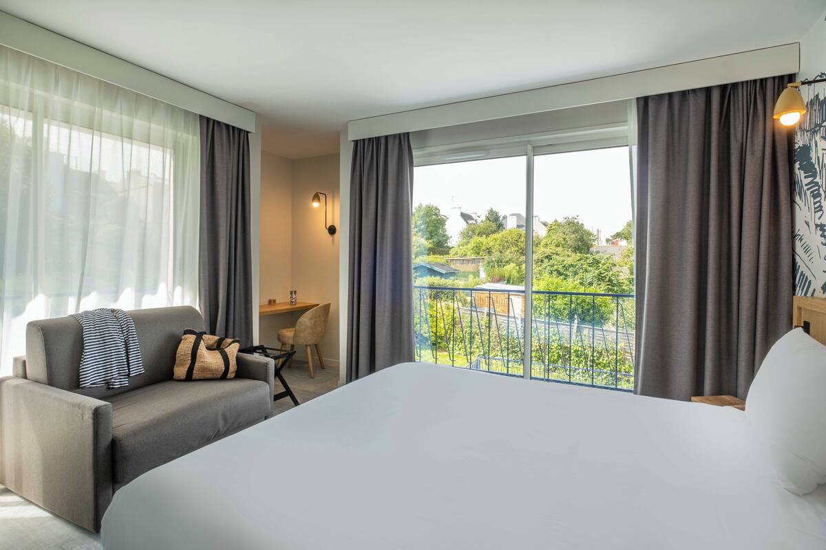 Ein Hotelzimmer mit einem großen Bett, einer Couch, einem Schreibtisch und einem Balkon mit Blick auf Grünflächen.
