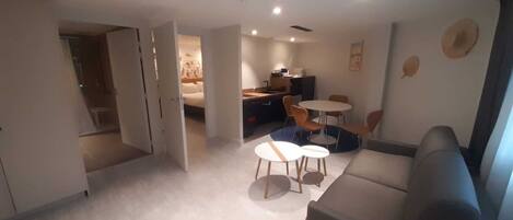A modern living room with a sofa, a dining table with chairs, and a kitchen area visible through an open door.