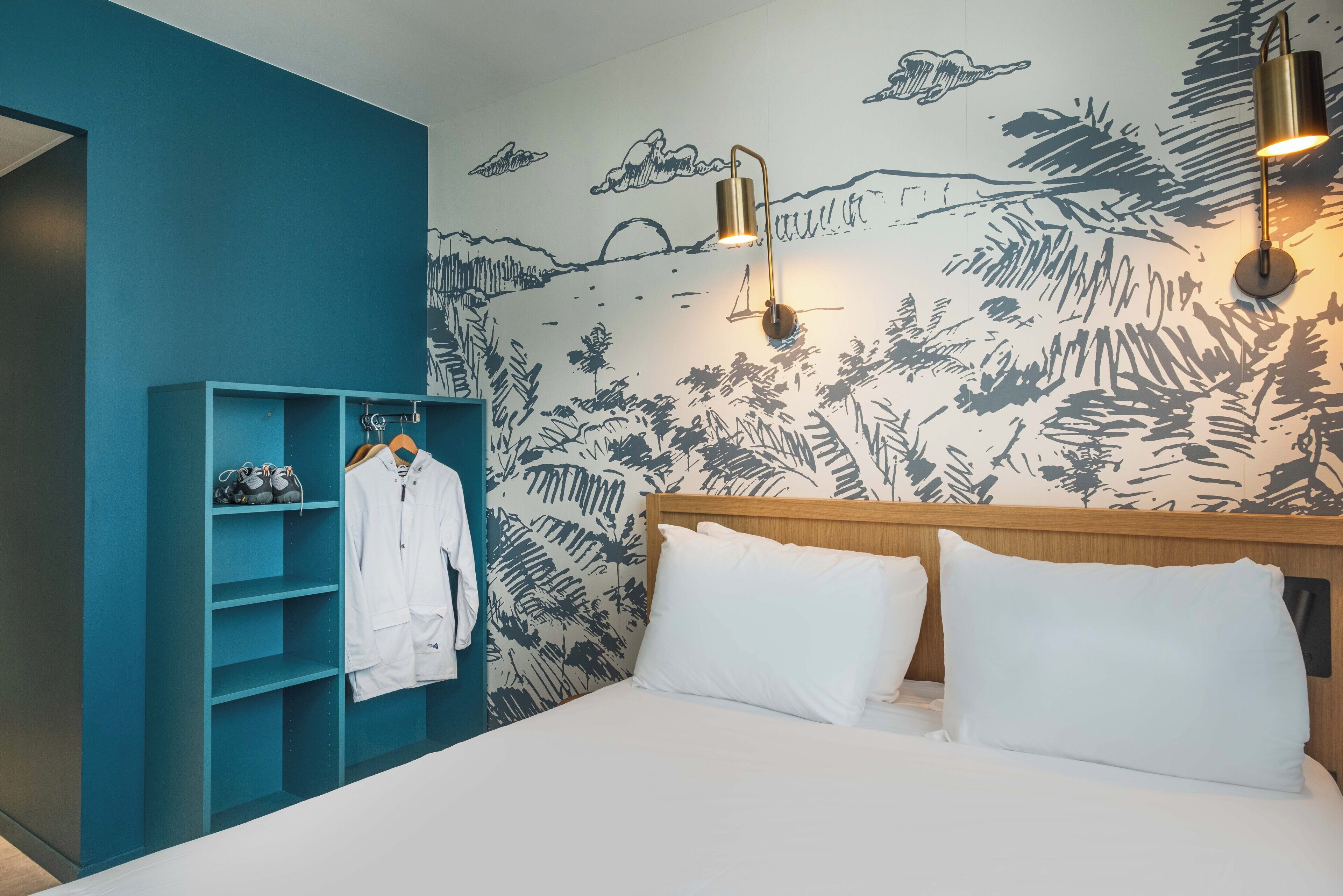 Une chambre à coucher avec un lit, des oreillers blancs, une tête de lit en bois, une armoire bleue et un mur orné d’une fresque sur le thème de la nature.