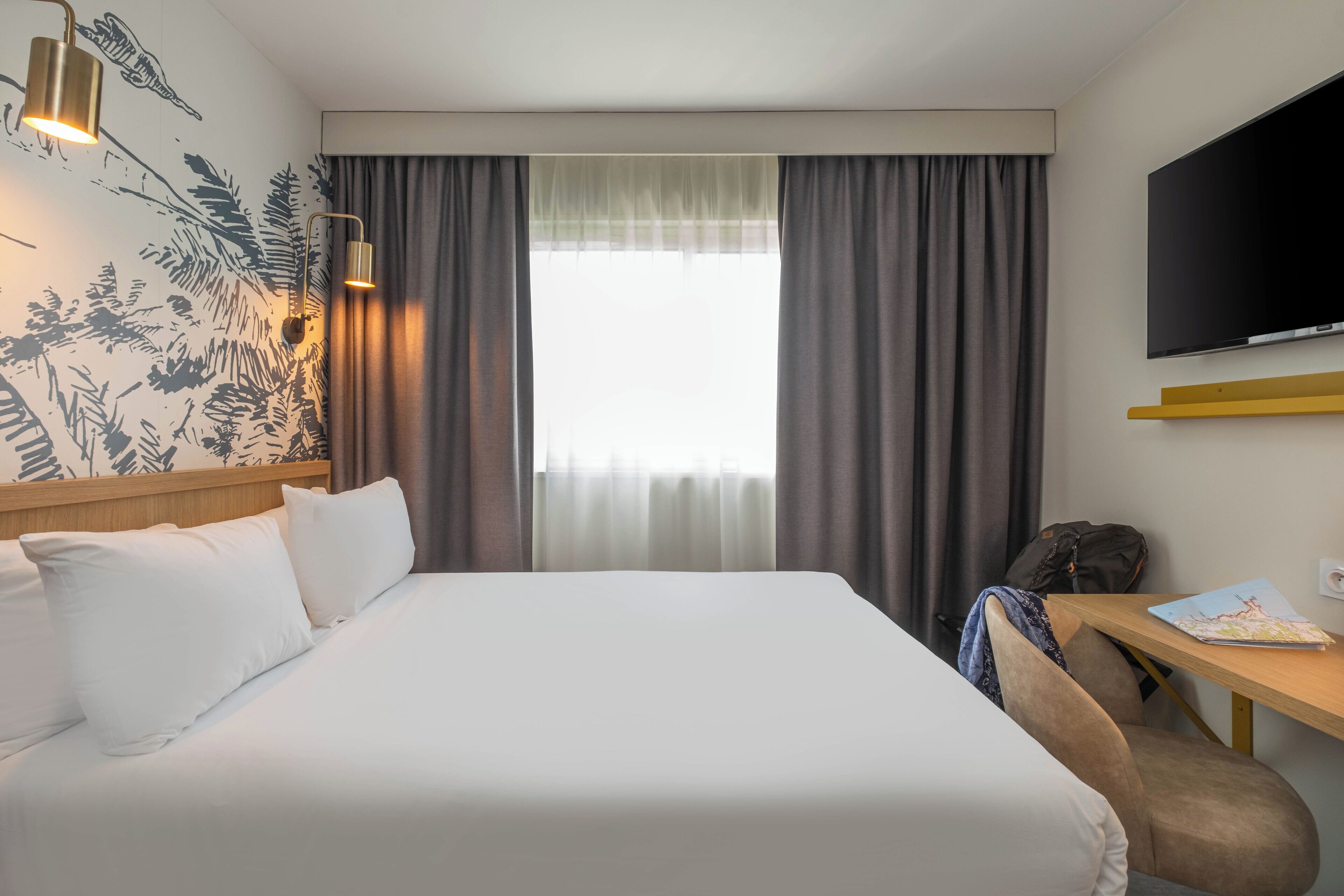 Quarto de hotel com cama, escrivaninha, cadeira, televisão e janela com cortinas.