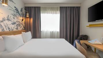 Quarto de hotel com cama, escrivaninha, cadeira, televisão e janela com cortinas.