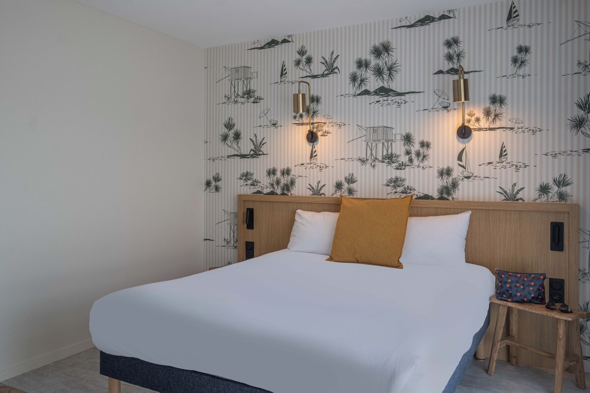 A hotel room with a large bed, a wooden headboard, and a wall with a tropical wallpaper design.