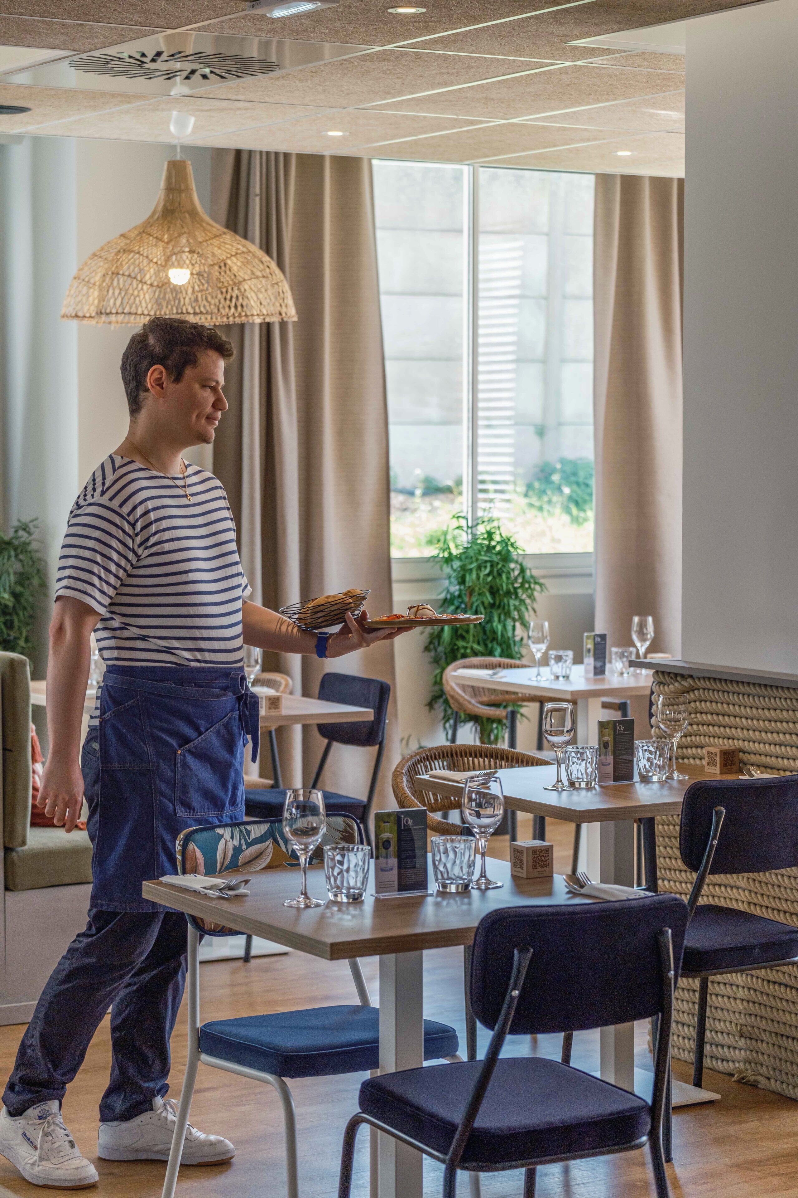 A waiter carrying a tray with food and drinks in a modern dining area with tables, chairs, and a large window.