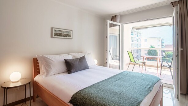 A hotel room with a bed, a bedside table with a lamp, a balcony with chairs, and a window with curtains.