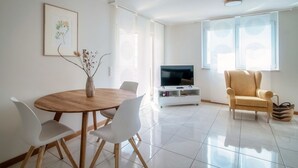 A modern living room with a wooden dining table, white chairs, a TV on a white stand, a beige armchair, and a large window with blinds.