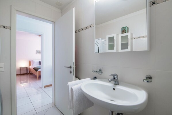 A modern bathroom with a white sink, mirror, and cabinets. A towel hangs on the door, and a bed is visible in the adjacent room.