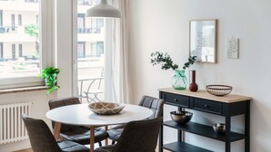 A modern dining area with a round table, dark chairs, a hanging light fixture, a window with curtains, and a wall-mounted shelf with decorative items. - Central two-room apartment with balcony (LOCARNO CITY 2) (Locarno)