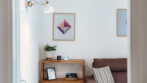 A room with a brown sofa, a wooden side table with a plant, a framed picture, and a wall-mounted lamp. - Central two-room apartment with balcony (LOCARNO CITY 2) (Locarno)