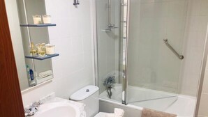 Une salle de bain équipée d’un lavabo, de toilettes et d’une douche. On peut voir des étagères où sont rangés les produits de toilette.