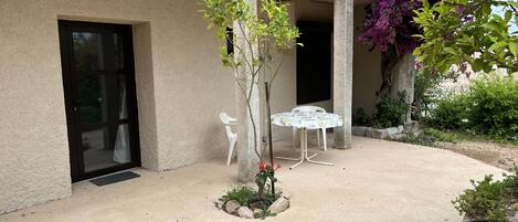 A courtyard with a concrete floor, a small tree, a white plastic chair, and a table with a patterned tablecloth.