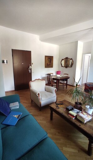 Ein Wohnzimmer mit einem türkisfarbenen Sofa, einem Holztisch mit Büchern, einem gemusterten Sessel, einem Esstisch mit Stühlen und einer Tür mit Schlüssel.