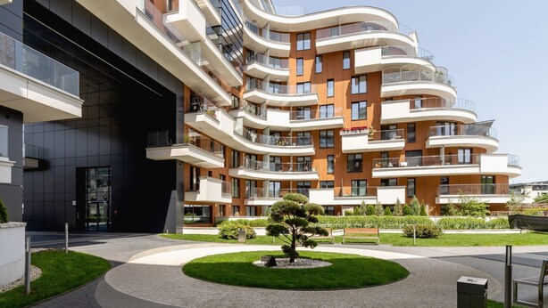 Un immeuble résidentiel moderne à plusieurs étages, doté de balcons, d’une cour aménagée avec un arbre et d’un chemin piétonnier pavé.