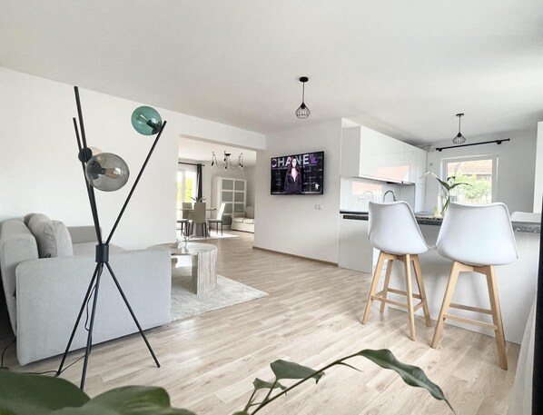A modern living room with a sofa, a coffee table, and a dining area with two chairs. A TV is mounted on the wall displaying a magazine cover.