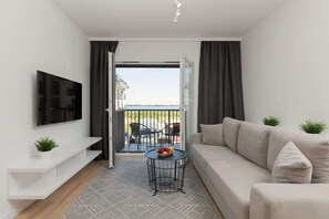 A modern living room with a sofa, a TV mounted on the wall, a small table with fruit, and a balcony with chairs.