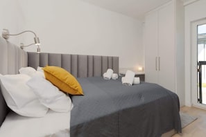 Une chambre moderne avec une tête de lit rembourrée de couleur grise, une couvre-lit grise, des oreillers blancs et un oreiller décoratif jaune. On peut voir des serviettes pliées sur le lit et une table de chevet avec une lampe.