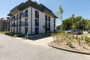 A modern multi-story residential building with balconies, a paved driveway, and a landscaped area with young trees.