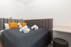 Une chambre d’hôtel avec une tête de lit recouverte d’un tissu gris, un lit avec une couvre-lit gris foncé, deux oreillers blancs, dont un avec un motif jaune moutarde, et deux serviettes enroulées posées sur le lit. On y trouve une table de chevet blanche et un petit tabouret noir.