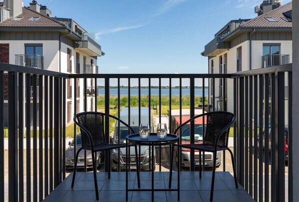 Un balcon avec deux chaises et une table, donnant sur une étendue d’eau et des voitures garées.
