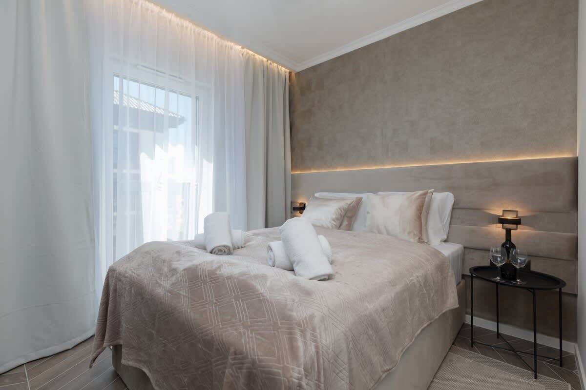 Ein modernes Hotelzimmer mit einem großen Bett, einem Nachttisch mit Lampe und Blick ins Freie durch leichte Vorhänge.