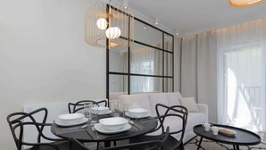 Ein moderner Essbereich mit einem schwarzen Tisch, gedeckt für vier Personen, umgeben von schwarzen Stühlen. Ein weißes Sofa und ein kleiner runder Tisch mit Tablett und Tassen stehen daneben. Ein großer Spiegel spiegelt den Raum wider, und eine außergewöhnliche Pendelleuchte hängt über dem Tisch.
